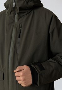 Oliivinvihreä vedenpitävä takki, jossa on vetoketjukiinnitys, säädettävä huppu ja mustat yksityiskohdat. Siinä on rintatasku ja kootut hihansuut eristämään lämpöä.