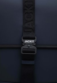 Dettaglio di uno zaino navy con una tracolla testurizzata con il marchio "JACK1T", una fibbia nera e una finitura in materiale liscio.