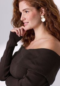 Brauner, gerippter Pullover mit schulterfreiem Ausschnitt und langen Ärmeln, der eine strukturierte Strickoptik aufweist. Das Modell trägt runde gold-weiße Ohrringe und präsentiert einen lässigen Look.