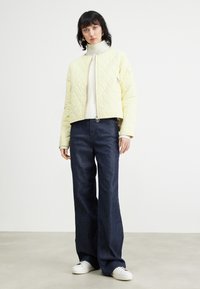 Gepatchte gelbe cropped Jacke, weißer Rollkragenpullover, dunkle weite Jeans und weiße Sneakers; auffälliges diagonales Muster auf der Jacke.