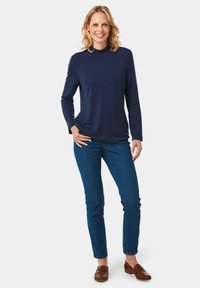 Marineblaues Langarmoberteil mit Rundhalsausschnitt, kombiniert mit eng anliegenden blauen Jeans und braunen Mokkas, stehend vor einem schlichten weißen Hintergrund.