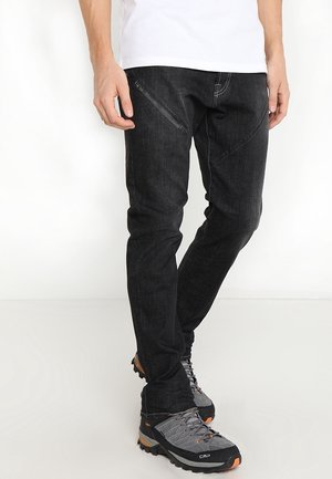Homme portant un pantalon noir slim fit avec une poche zippée diagonale, chaussures de randonnée grises et orange, debout devant un fond blanc.
