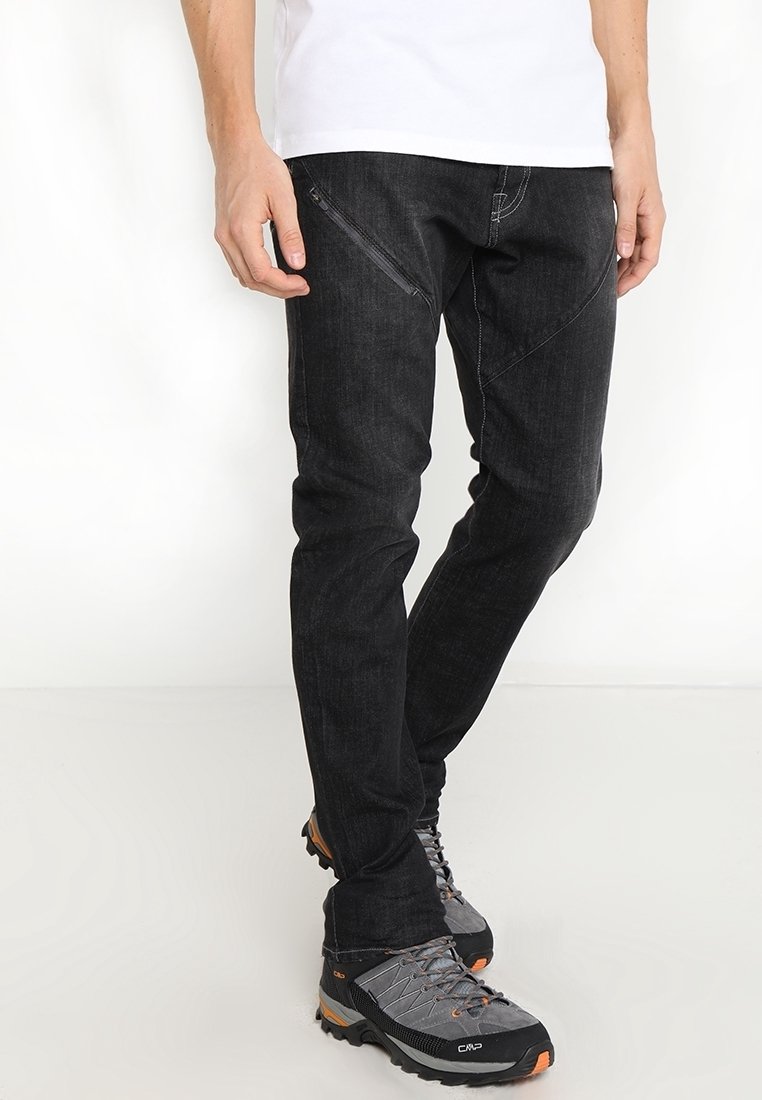 Homme portant un pantalon noir slim fit avec une poche zippée diagonale, chaussures de randonnée grises et orange, debout devant un fond blanc.