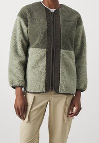 Veste en polaire verte à surface texturée, avec fermeture éclair et accents marron foncé. Manches longues et design de col arrondi.