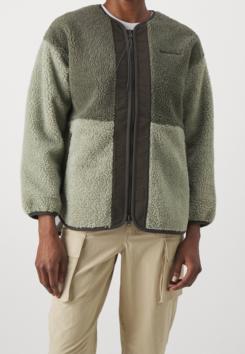 Veste en polaire verte à surface texturée, avec fermeture éclair et accents marron foncé. Manches longues et design de col arrondi.