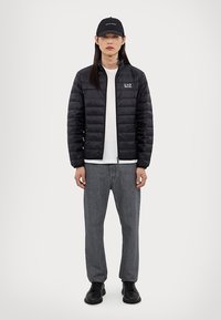 Doudoune noire avec motif matelassé et fermeture éclair, associée à un t-shirt blanc, un jean gris et des baskets noires. Casquette noire incluse.