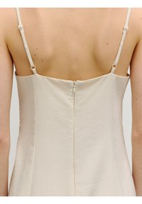 Ecrufarbenes Kleid mit dünnen Trägern, glatter Textur und verdecktem Reißverschluss auf der Rückseite. Es hat ein tailliertes Oberteil und ein ausgestelltes Rockdesign.