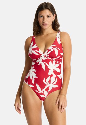 Mujer con un traje de baño rojo de una pieza con grandes estampados florales blancos, de pie con los brazos relajados sobre un fondo liso.