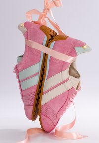 Paire de baskets en daim rose avec des rayures blanches et bleu clair, semelles marron, attachées ensemble par un ruban rose en satin.