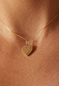 Collier en or avec un pendentif en forme de cœur au centre lisse et une bordure de petites pierres claires. Fond en peau texturée.