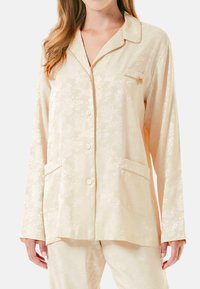 Conjunto de pijama de color beige con un patrón floral, de mangas largas, con botones en la parte delantera y dos bolsillos frontales. La tela tiene una textura suave y lisa.