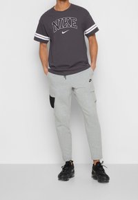 Šedé tepláky Nike s černými akcenty na kapsách a šedým stahovacím páskem. Šedé tričko s velkým černým logem "Nike" a bílými pruhy.