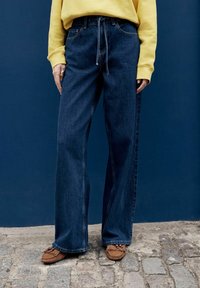 Persona valkājot tumši zilas plataji džinsus, dzeltenu sporta kreklu un brūnas mokasīnu kurpes, stāv uz bruģa pret zilu sienu.