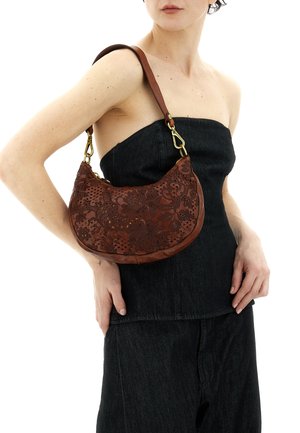 Donna che indossa un top nero senza spalline e pantaloni, con una borsa a tracolla in pelle marrone con motivi floreali in rilievo e dettagli dorati.