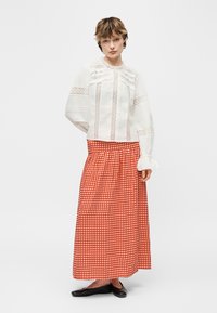 Vrouw met kort haar, gekleed in een witte kanten blouse met opgeblazen mouwen en een lange rood-wit geruite rok met zwarte platte schoenen.