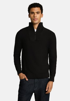 Junger Mann mit lockigem Haar, der einen schwarzen Pullover mit halbem Reißverschluss und dunkle Jeans trägt, steht mit einer Hand in der Tasche vor einem schlichten Hintergrund.