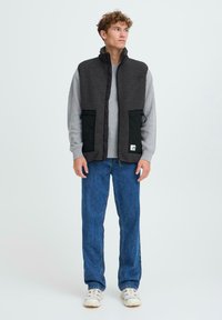 Grau gefütterte Weste mit schwarzen Stofftaschen, hohem Kragen und Reißverschluss, getragen über einem hellgrauen Hemd und kombiniert mit blauen Jeans.