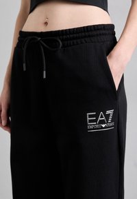 Čierne bavlnené tepláky s elastickým pásom, šnúrkou na uväzovanie, bočnými vreckami a strieborným vyšívaným logom EA7 Emporio Armani.