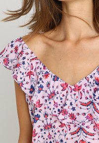 Femme portant une robe rose à fleurs avec des manches à volants dénudées, dévoilant le cou et la zone des clavicules.