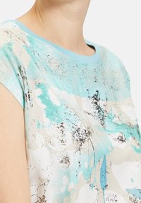 Primer plano de una persona que lleva una camisa de manga corta con un patrón abstracto en azul, blanco y beige, y un escote azul claro.