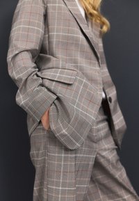 Grey plaid suit featuring a checkered pattern in black and red accents. Tailored fit with noticeable structured shoulders and pockets.