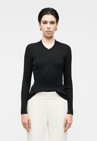 Camisola preta de malha canelada com decote em V e mangas compridas, apresentando um corte justo e bainha divisória. Usada sobre calças de cor clara.