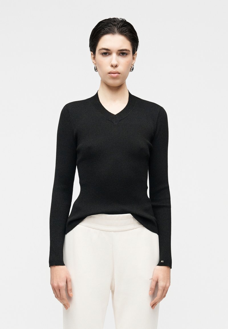 Černý žebrovaný pletený svetr s výstřihem do V a dlouhými rukávy, s přiléhavým střihem a rozparky na lemu. Nošený přes světlé kalhoty.