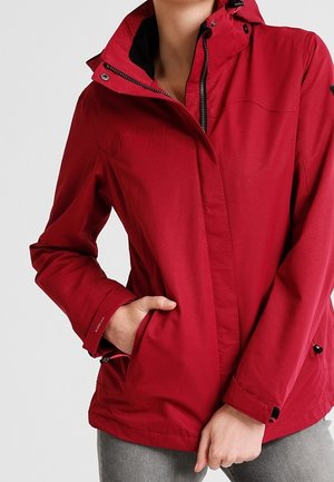 Personne portant une veste rouge à capuche avec les mains dans les poches, debout devant un fond clair uni.