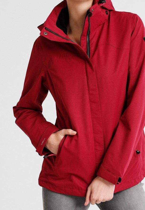 Personne portant une veste rouge à capuche avec les mains dans les poches, debout devant un fond clair uni.