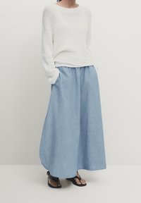 Donna che indossa un maglione bianco a maglia larga, pantaloni a gamba larga blu chiaro e sandali neri, in piedi con le mani nelle tasche su uno sfondo semplice.