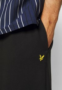 Pantaloni sportivi neri con una texture liscia presentano un piccolo logo giallo di un'aquila sulla tasca. La parte superiore mostra una maglietta a righe blu navy.