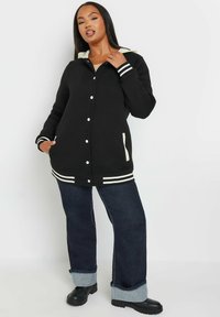 Schwarze, lange Hoodie-Jacke mit weißen Akzenten, vorderen Knöpfen und zwei Seitentaschen. Kombiniert mit blauen Jeans mit umgeschlagenen Säumen und schwarzen Stiefeln.