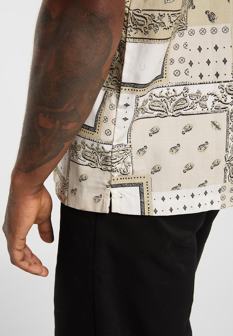 Chemise à manches courtes en tissu beige à motifs avec accents noirs, dotée d'une coupe décontractée et de fentes latérales près de l'ourlet. Pantalon noir présenté en dessous.