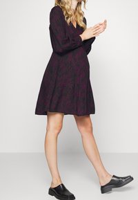 Czarna sukienka z teksturowanym fioletowym wzorem fal, dekoltem w kształcie litery V, długimi rękawami i rozkloszowanym dołem, zestawiona z czarnymi butami wsuwanymi.