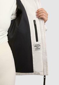 Giubbotto imbottito con strato esterno bianco e fodera nera. Presenta una tasca interna con zip e un'etichetta in tessuto con scritta.