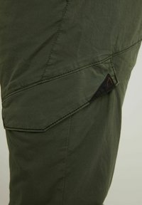 Primer plano de pantalones de tela verde oliva con una solapa de bolsillo asegurada por un pequeño parche negro triangular con costuras naranjas.