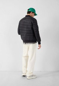 Chaqueta negra acolchada con costuras horizontales, puños y cuello acanalados, combinada con pantalones amplios color crema y una gorra verde. Look deportivo casual.