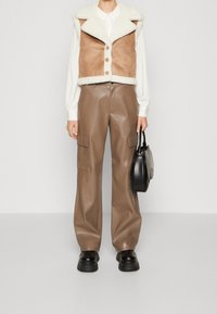 Brun fårskinnsväst med krämfärgad krage och knappar, kombinerad med högmidjade bruna läderbyxor och svarta kraftiga skor, hållande en svart väska.