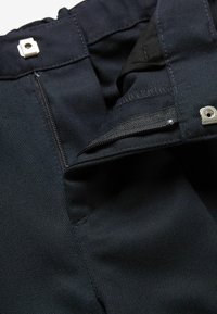 Pantalons en tissu bleu marine avec une braguette zippée, fermeture à pression argentée, et des détails de couture visibles, mettant en valeur une surface texturée.