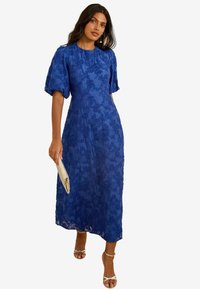 Sininen kukkakuvioinen pitsimidi-mekko, jossa on lyhyet puffihihat, pyöreä pääntie, teksturoitu kangas, vartalonmyötäinen vyötärö. Mekko on yhdistetty hopeiseen clutch-laukkuun ja sanka-sandaaleihin.