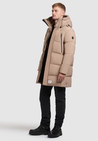 Bărbat îmbrăcat într-o geacă puffer cu glugă beige, cu buzunare cu fermoar, pantaloni negri și cizme negre, stând de profil pe un fundal gri deschis.