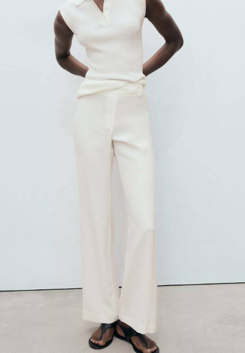Top senza maniche di colore crema lavorato a maglia con colletto, abbinato a pantaloni larghi crema; modella che indossa sandali neri, design semplice.