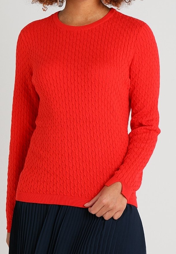 Femme portant un pull en maille texturée rouge vif à manches longues et col rond, associé à une jupe plissée foncée.