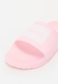 Rózsaszín csúszó szandál széles pánttal, melyen a "HUGO" felirat található. Simaszövet, lapos talp és enyhén lekerekített szélek jellemzik.