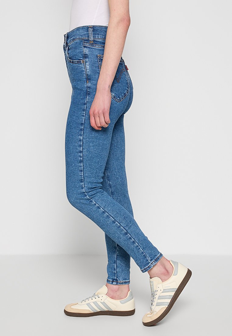 Jeans skinny azules hechos de tela de mezclilla, con una cintura alta, un diseño ajustado y estilo tradicional de cinco bolsillos. Combinados con zapatillas ligeras.