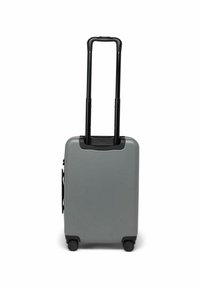 Valise rigide grise avec une poignée noire rétractable et quatre roues noires. Elle présente un design épuré et une poche latérale.