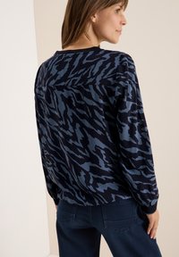 Långärmad topp i marinblått med ett blått abstrakt mönster. Har rundad halsringning och lös passform. Matchas med mörkblå jeans.