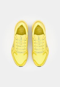 Zapatillas deportivas amarillas con una parte superior de malla texturizada, acentos reflectantes y una suela acolchada. Cuenta con cordones planos y una marca impresa en la lengüeta.