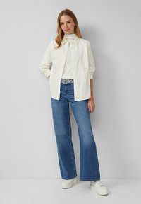 Witte knop-overhemd over een wit topje, gecombineerd met wijdvallende blauwe jeans en witte sneakers. De outfit heeft een geavanceerde riem.