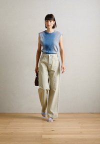 Haut sans manches à côtes bleu clair, pantalon large beige taille haute, et sandales bleu clair, tenant un petit sac à main marron, arrière-plan de sol en bois.